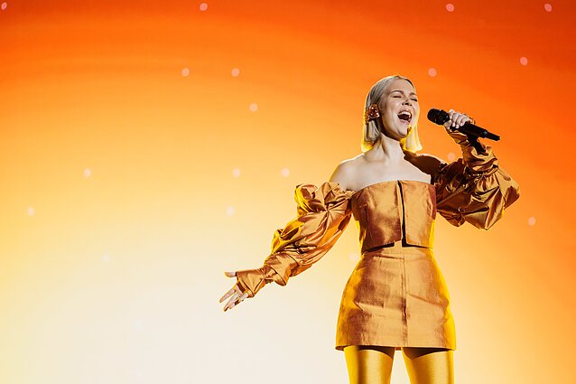 A woman singing into a microphone. She is wearing an off-the-shoulder minidress with matching tights and is singing on stage in front of a soft-lit backdrop