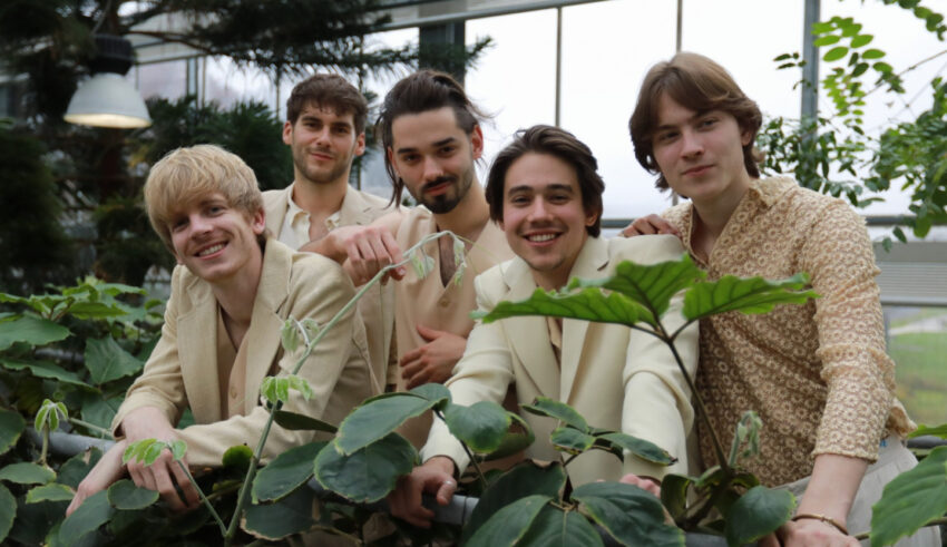 The five members of Joker Out in tan jackets and shirts, surrounded by green plants.