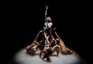 Raiven surrounded by male dancers against a black backdrop.